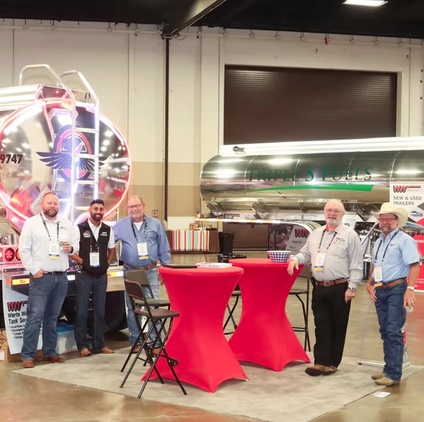 Five men standing and smiling near two red-covered tables at an indoor trade show booth with large fuel tanks and trailers displayed behind them.